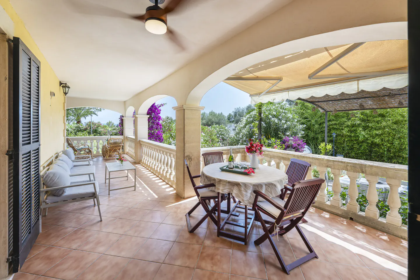 Covered patio with tile floor, arched openings, and white balustrade. Table with chairs, sofa, and lush green landscape in the background.