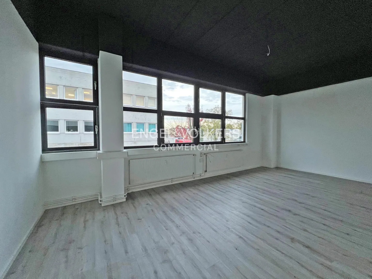 Empty commercial space with gray wood-look floors, white walls, black ceiling, and large windows. Radiators are under the windows.