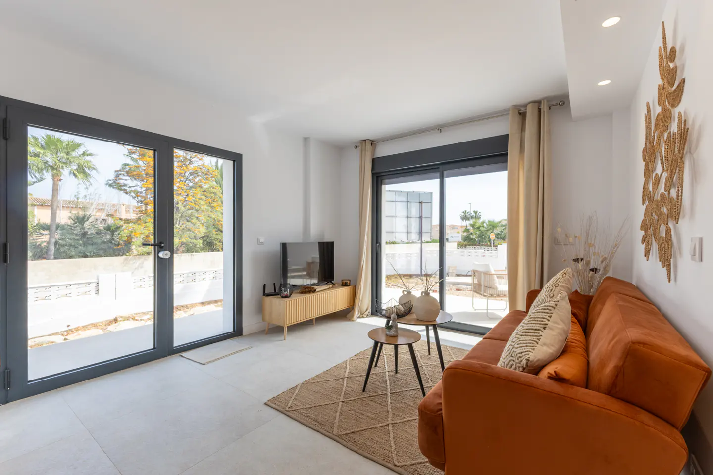 Bright living room with orange sofa, jute rug, and TV. Large windows offer outdoor views. Modern decor with neutral tones and natural textures.