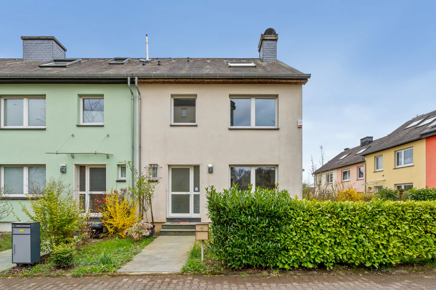 A two-story townhouse with a beige facade, white trim, and a green hedge in front. A green townhouse is on the left.