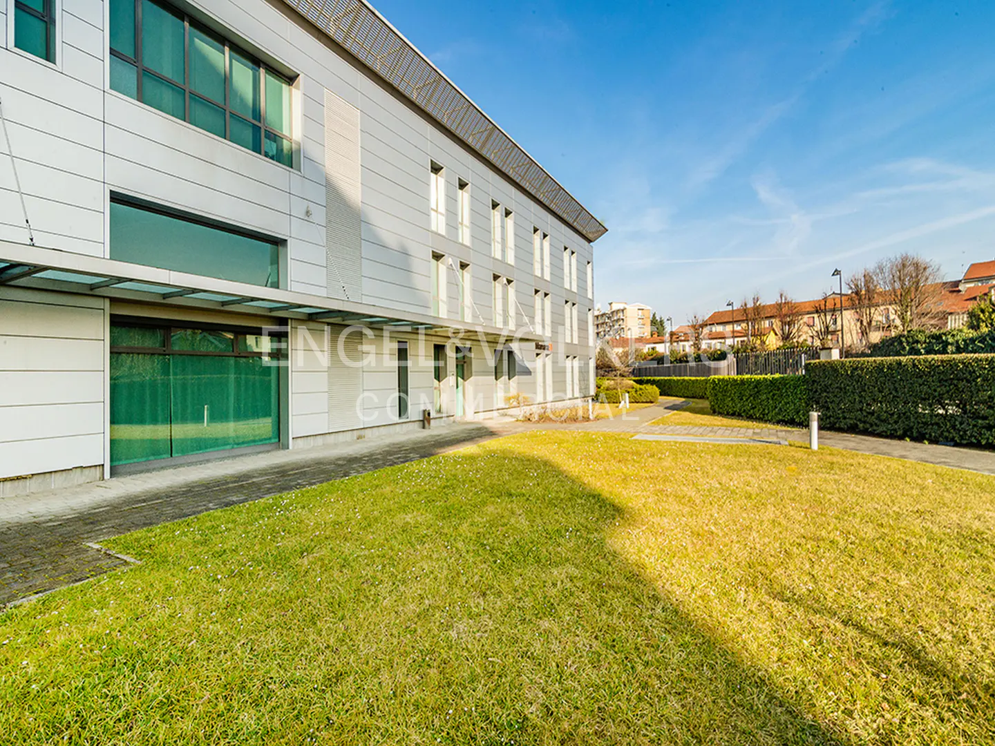 Modern office building with a white metal facade, large windows, and a green lawn under a clear blue sky.