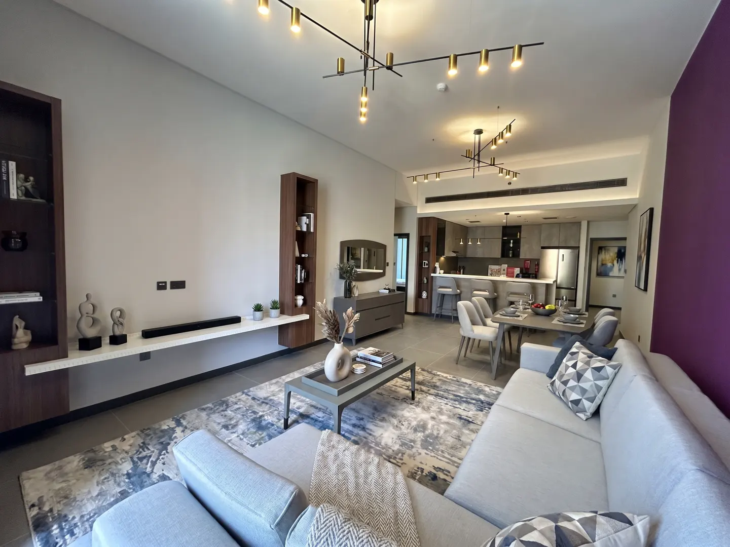 A modern apartment living room with a gray sofa, coffee table, and open kitchen. The walls are white and purple, with dark wood shelving.
