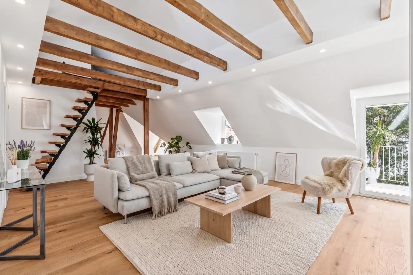 Bright living room with exposed wood beams, white walls, wood floors, a gray sectional sofa, and a staircase.