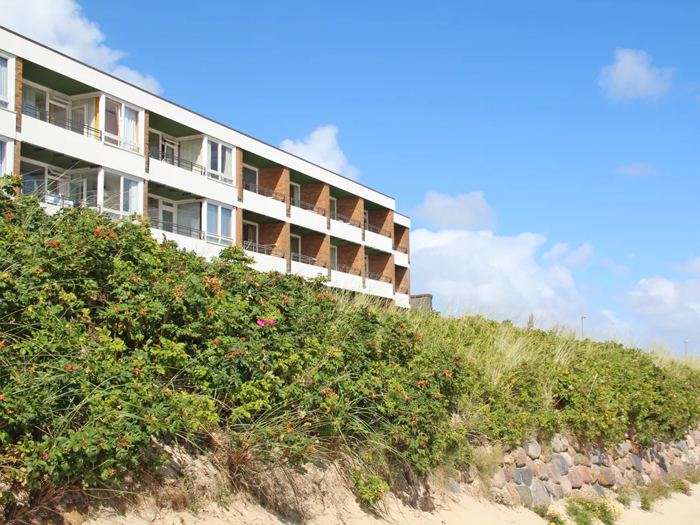 Beachfront property with a white building, balconies, and green bushes against a blue sky.