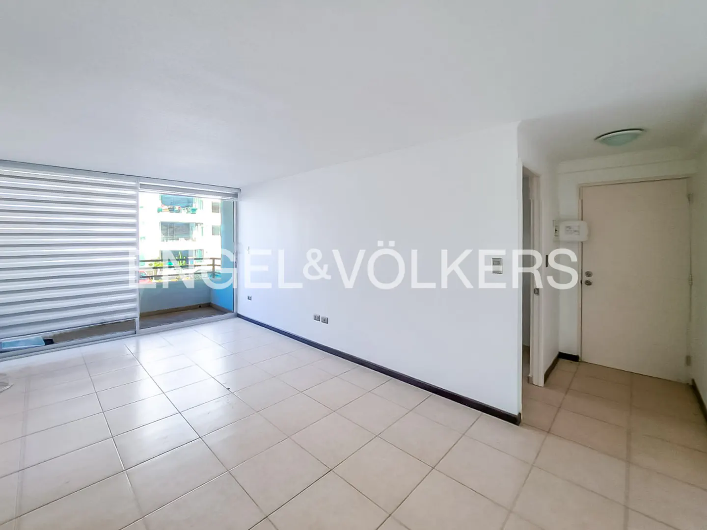 Empty room with white walls, tile floor, and balcony. Engel & Völkers logo is superimposed on the wall. A door is visible on the right.