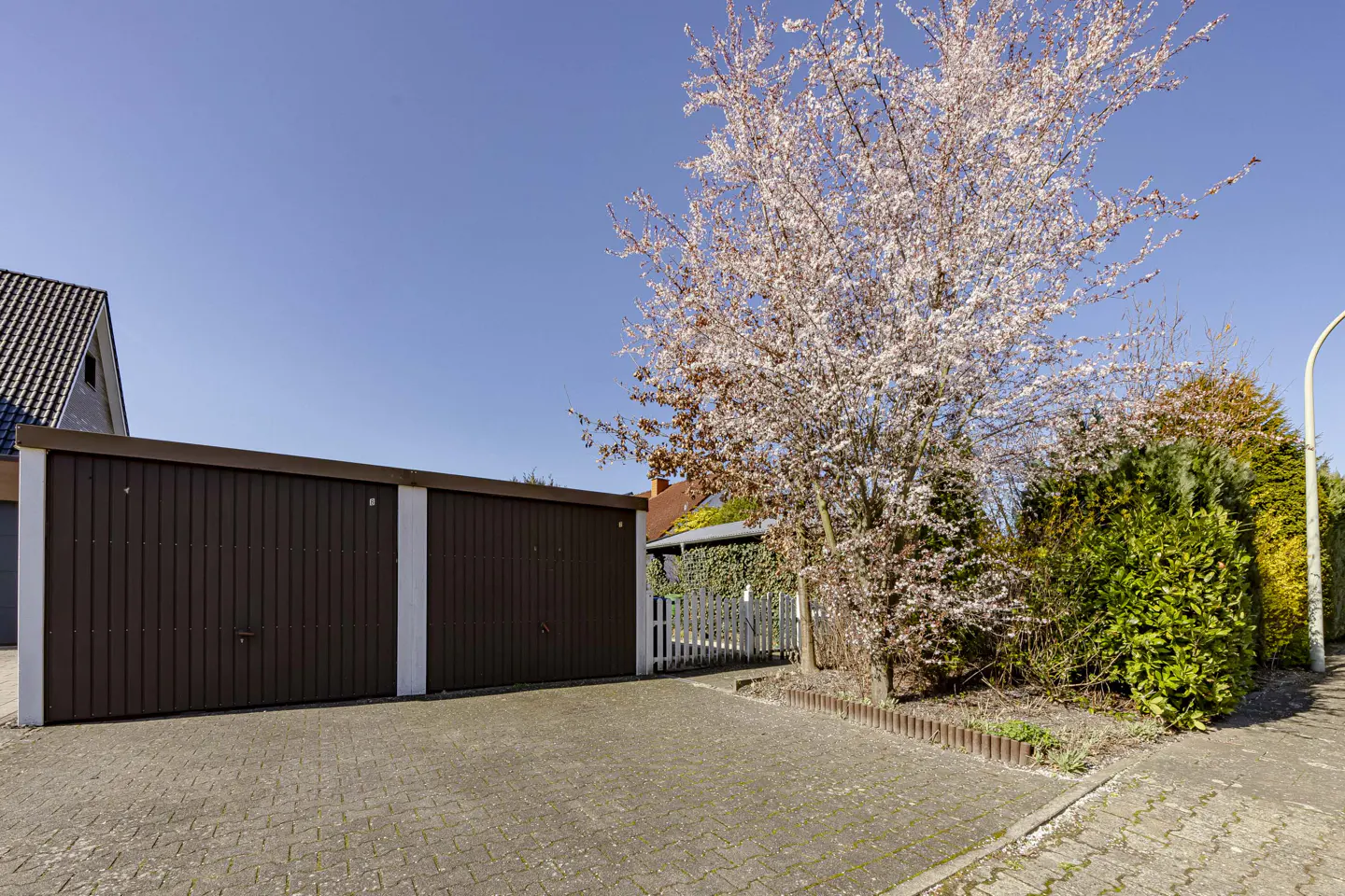 A brown two-car garage with a paved driveway, a flowering tree, and green bushes under a blue sky.