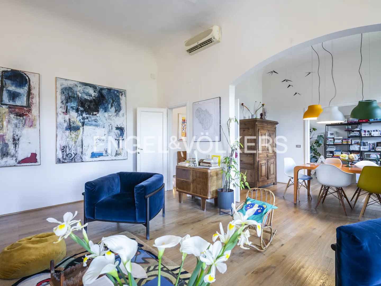Bright, open living space with hardwood floors, blue velvet chairs, and abstract art. Dining area visible through archway.