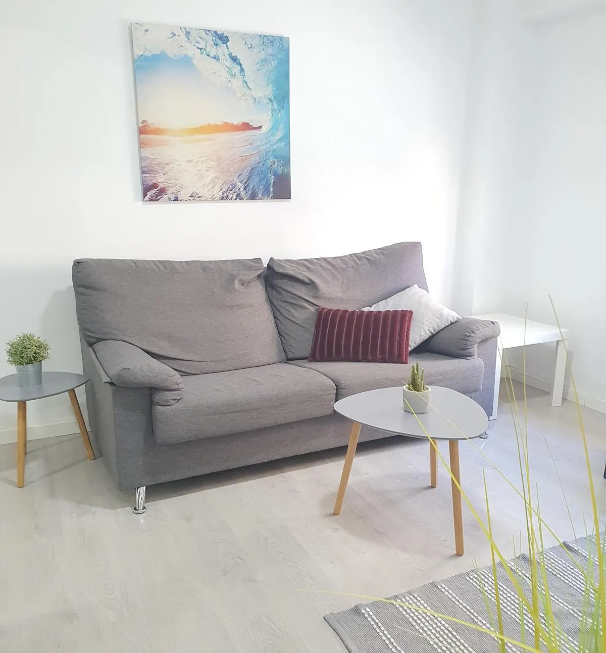 Modern living room with a grey sofa, a wave art print on the wall, two small coffee tables, and light wood-look flooring.