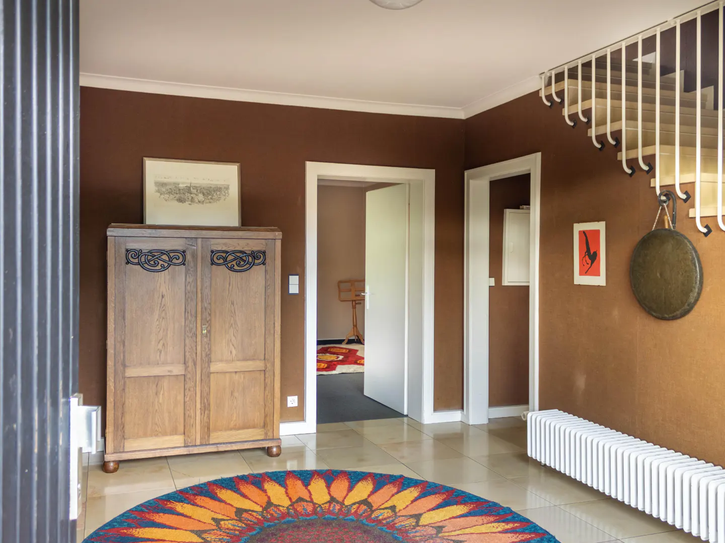 Foyer with brown walls, white trim, and a colorful round rug. A wooden cabinet sits near an open doorway. A gong hangs near a white radiator.