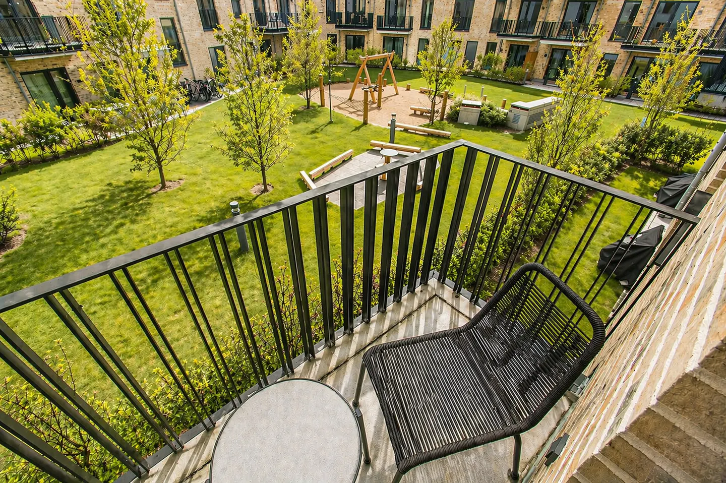 Balcony view of a green courtyard with playground. Black metal railing, chair, and small table on the balcony. Brick building in background.