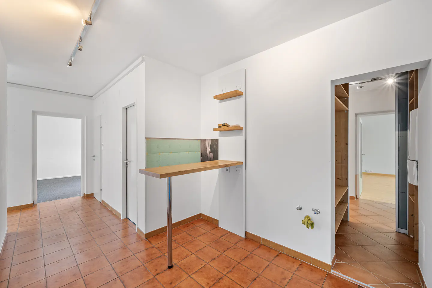 Bright hallway with terracotta tile floors, a wooden breakfast bar, wall shelves, and doorways leading to other rooms.