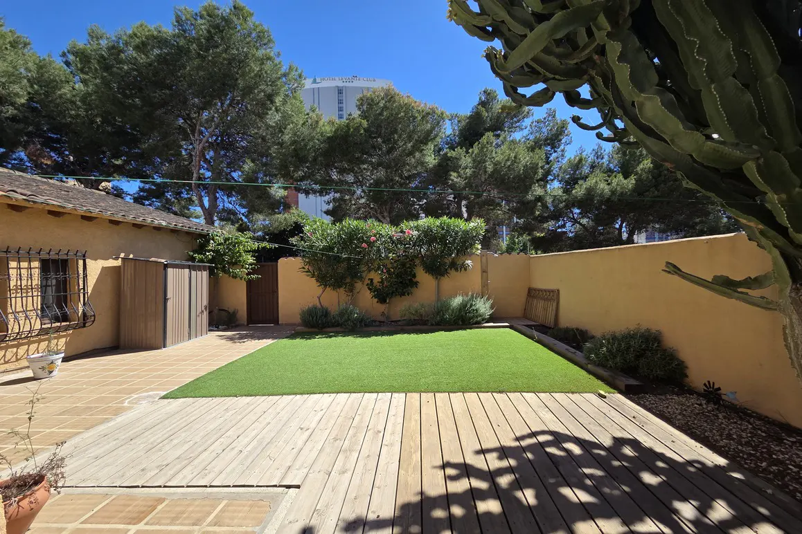 A backyard with a wooden deck, green grass, and yellow walls, surrounded by trees under a blue sky.