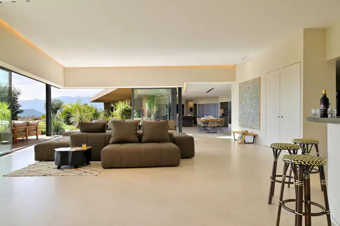 Open-concept living room with a brown sectional sofa, black coffee table, and floor-to-ceiling windows overlooking a lush green landscape.