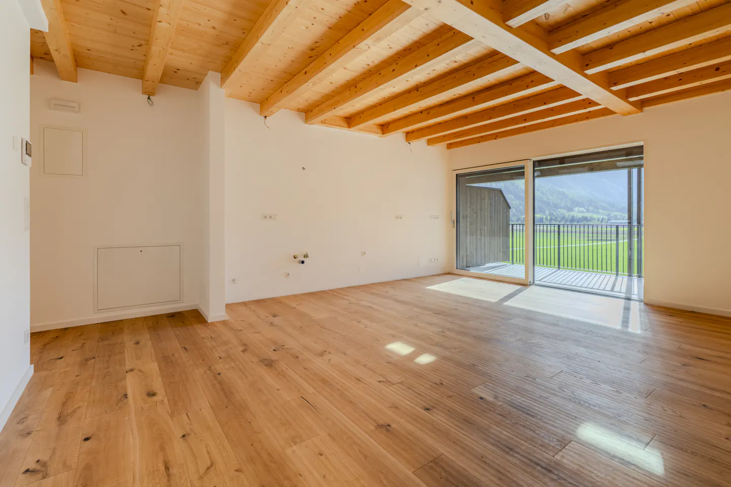 Bright, empty room with wood floors, white walls, and exposed wood beam ceiling. Sliding glass doors open to a balcony with a green field view.