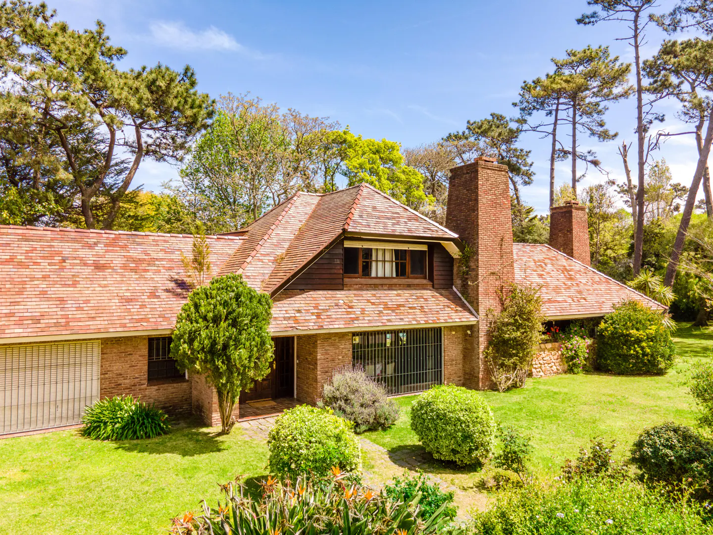 Exterior view of a brick house with a red tile roof, green lawn, and trees under a blue sky.