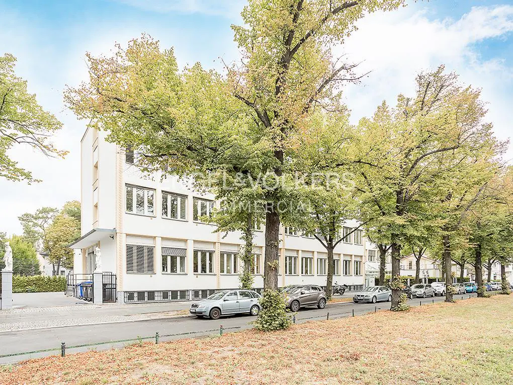 A white multi-story office building surrounded by large green trees with cars parked along the street in front.