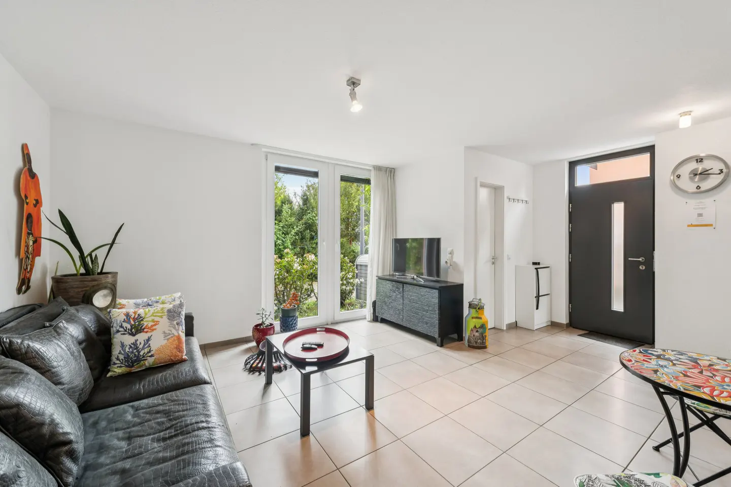 Bright living room with a black leather sofa, glass coffee table, TV on a black cabinet, and a dark front door.