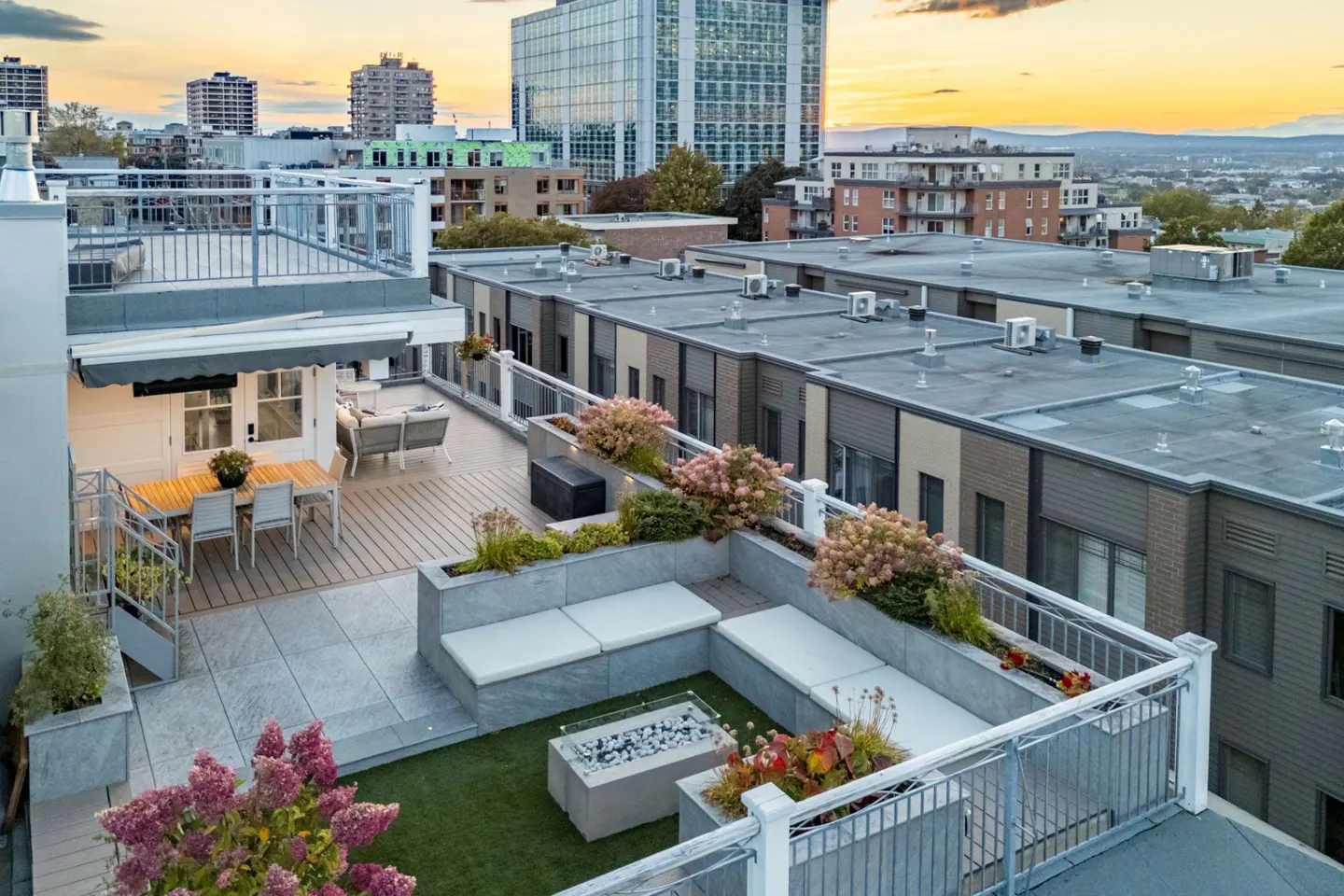Rooftop terrace at sunset with dining area, built-in seating, fire pit, and views of city buildings.