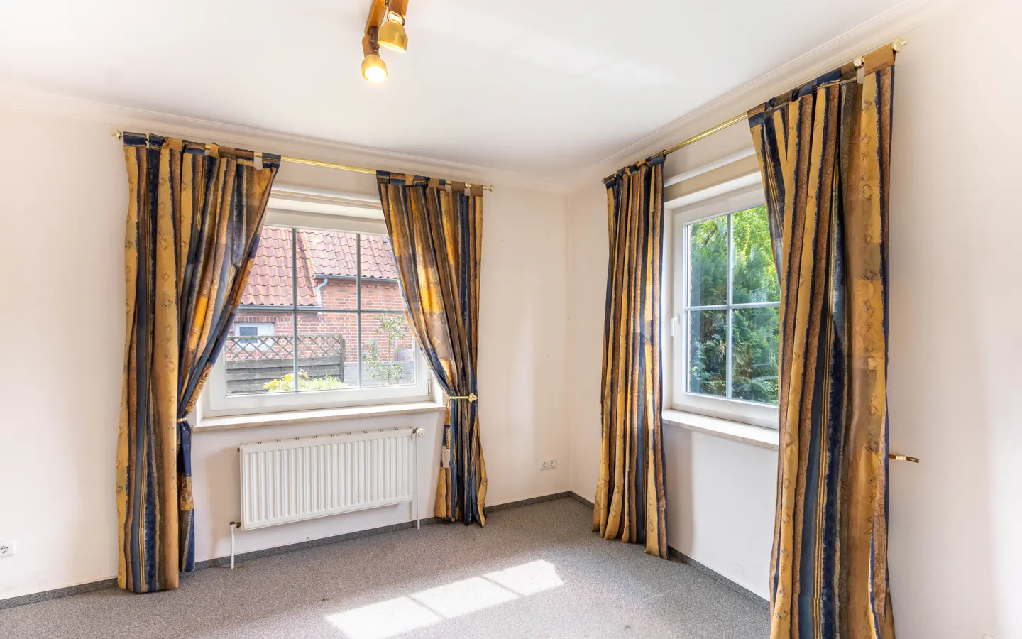 A bright, empty room with two windows dressed in gold and blue striped curtains. A radiator sits beneath one window.