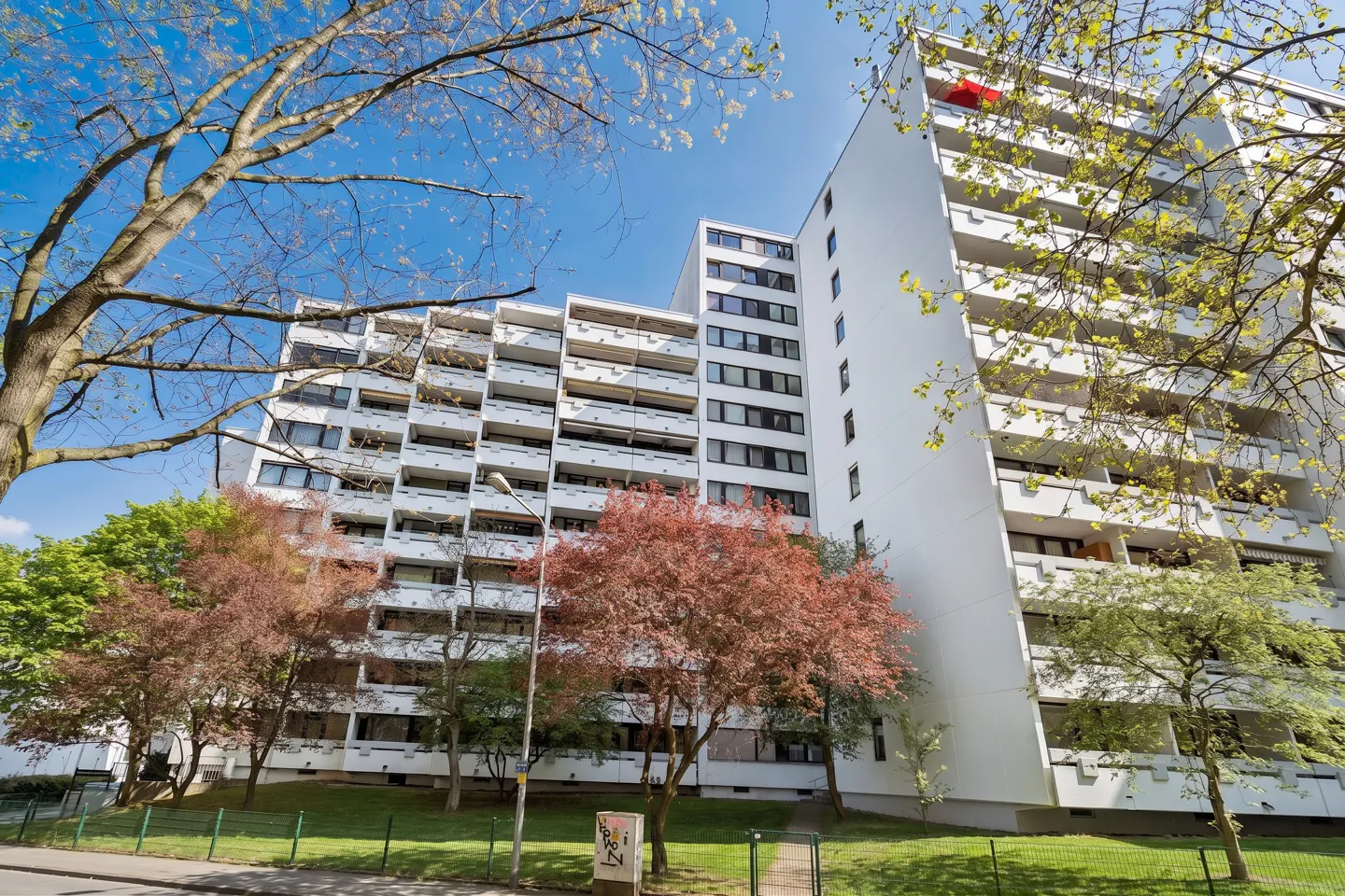 Exterior view of a tall, white apartment building with balconies, framed by trees and a blue sky.