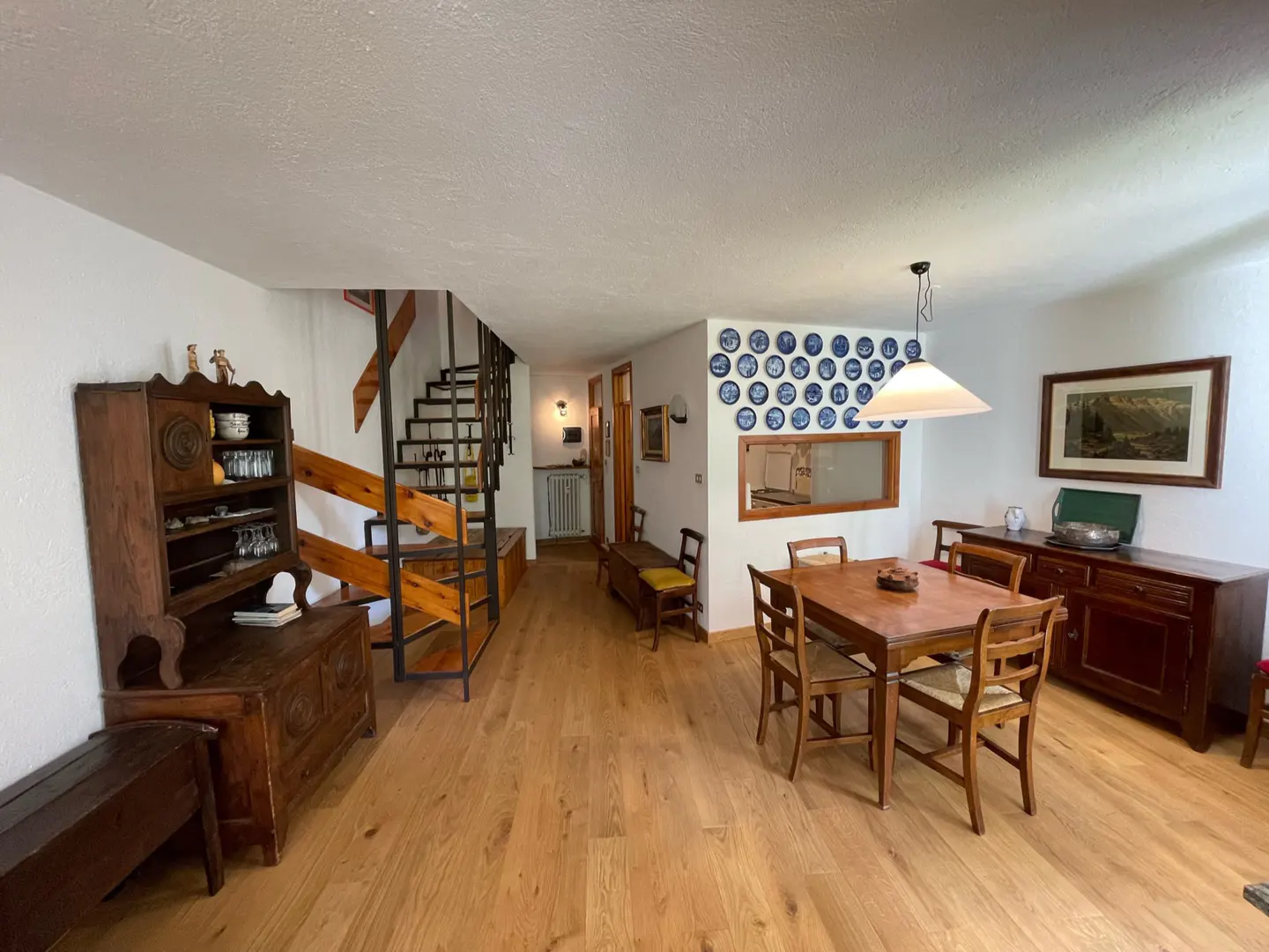 A bright, open-concept dining and living area with wooden floors, furniture, and a staircase. Blue decorative plates adorn the wall.