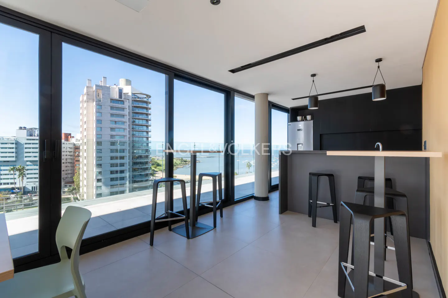 Modern kitchen with black bar stools, gray counter, and large windows overlooking a city and ocean view.