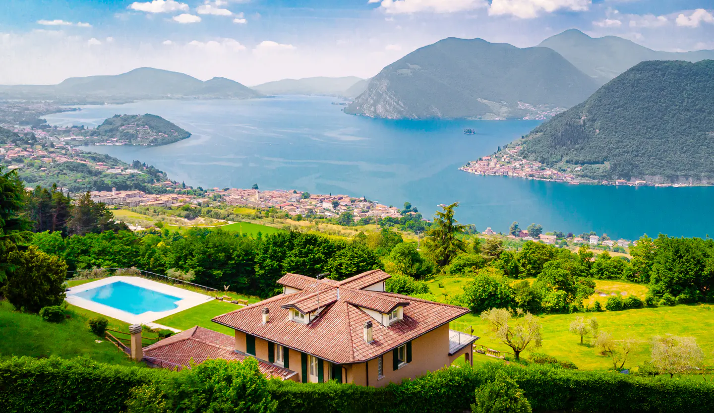Scenic view of a tan house with a red tile roof and a blue pool, overlooking a lake, town, and mountains.