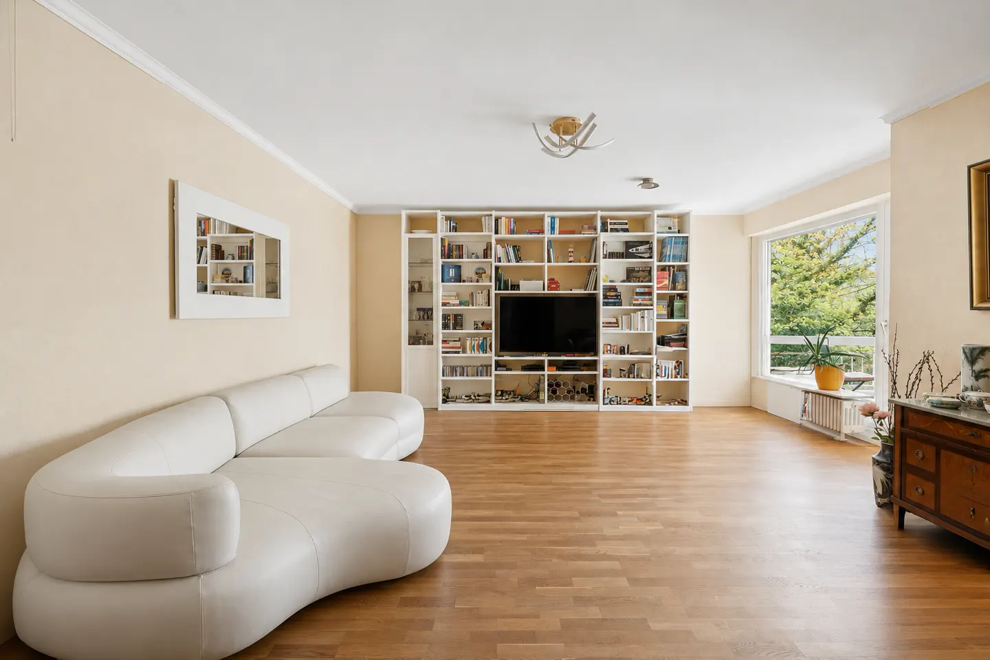 Bright living room with wood floors, a white curved sofa, and a large white bookcase with a TV in the center. A window shows green trees.