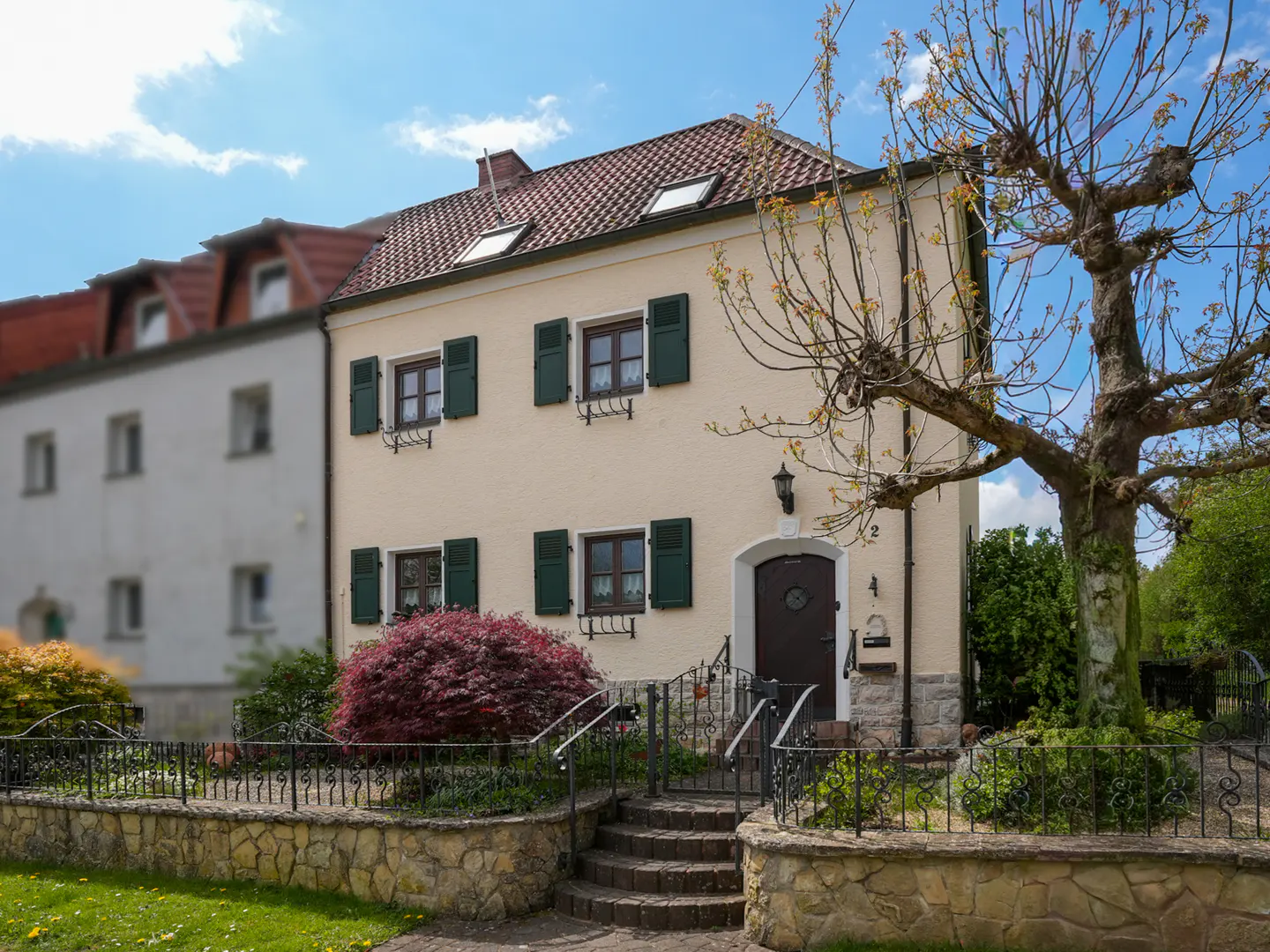 A cream-colored two-story house with dark green shutters, a tiled roof, and a stone front garden wall under a blue sky.