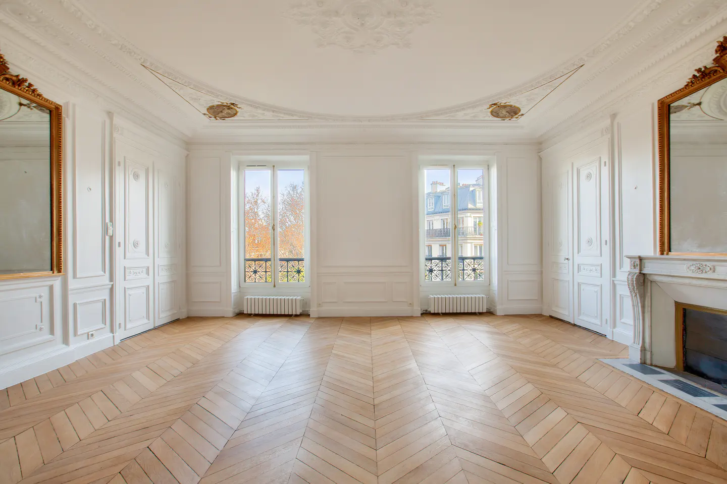 Bright, empty room with herringbone wood floors, white walls, two windows, and ornate gold-framed mirrors and fireplace.