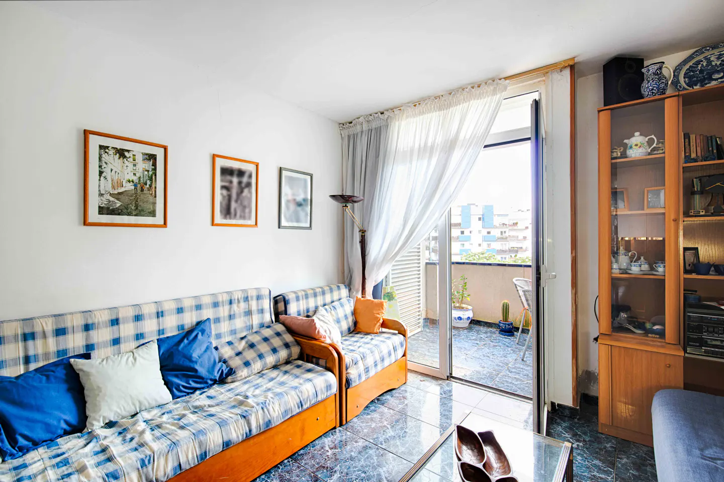 Bright living room with blue plaid sofas, white walls with framed art, and an open balcony with city views.