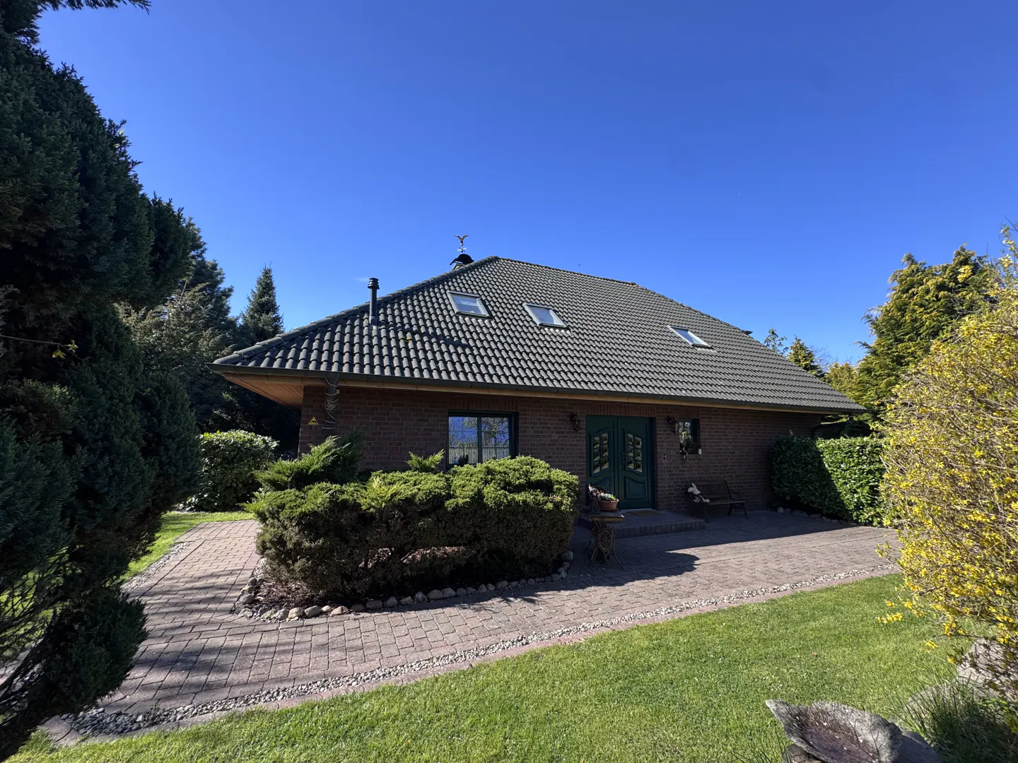 Brick house with a dark green tiled roof, skylights, and green double doors. A brick path leads to the entrance. Green lawn and trees surround the house.
