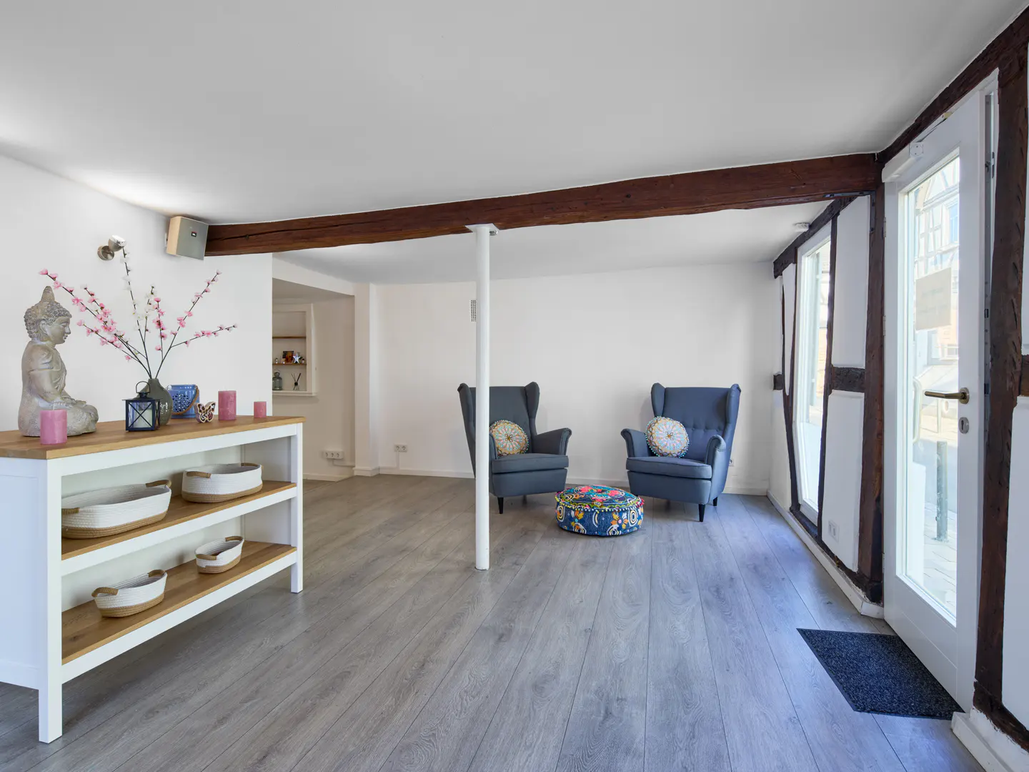 Bright living room with gray wood floors, white walls, and exposed beams. Two blue armchairs face a white door with dark wood trim. A white shelf holds a Buddha statue.