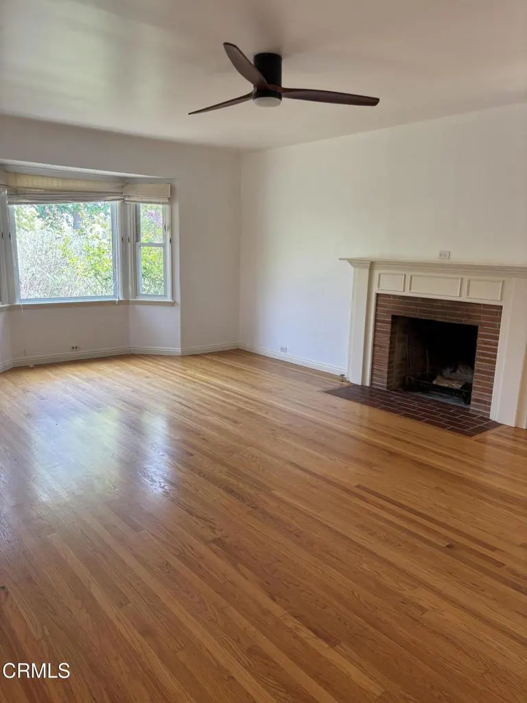 Empty living room with hardwood floors, a brick fireplace, large windows, and a ceiling fan.