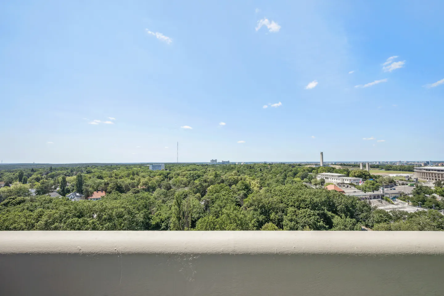 View from a balcony overlooking a green, tree-filled landscape under a blue sky with scattered clouds.