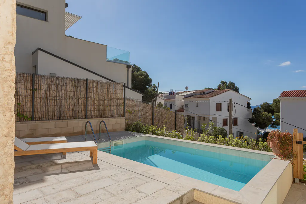 Outdoor pool with lounge chairs, metal ladder, and a bamboo fence. White buildings and blue sky in the background.