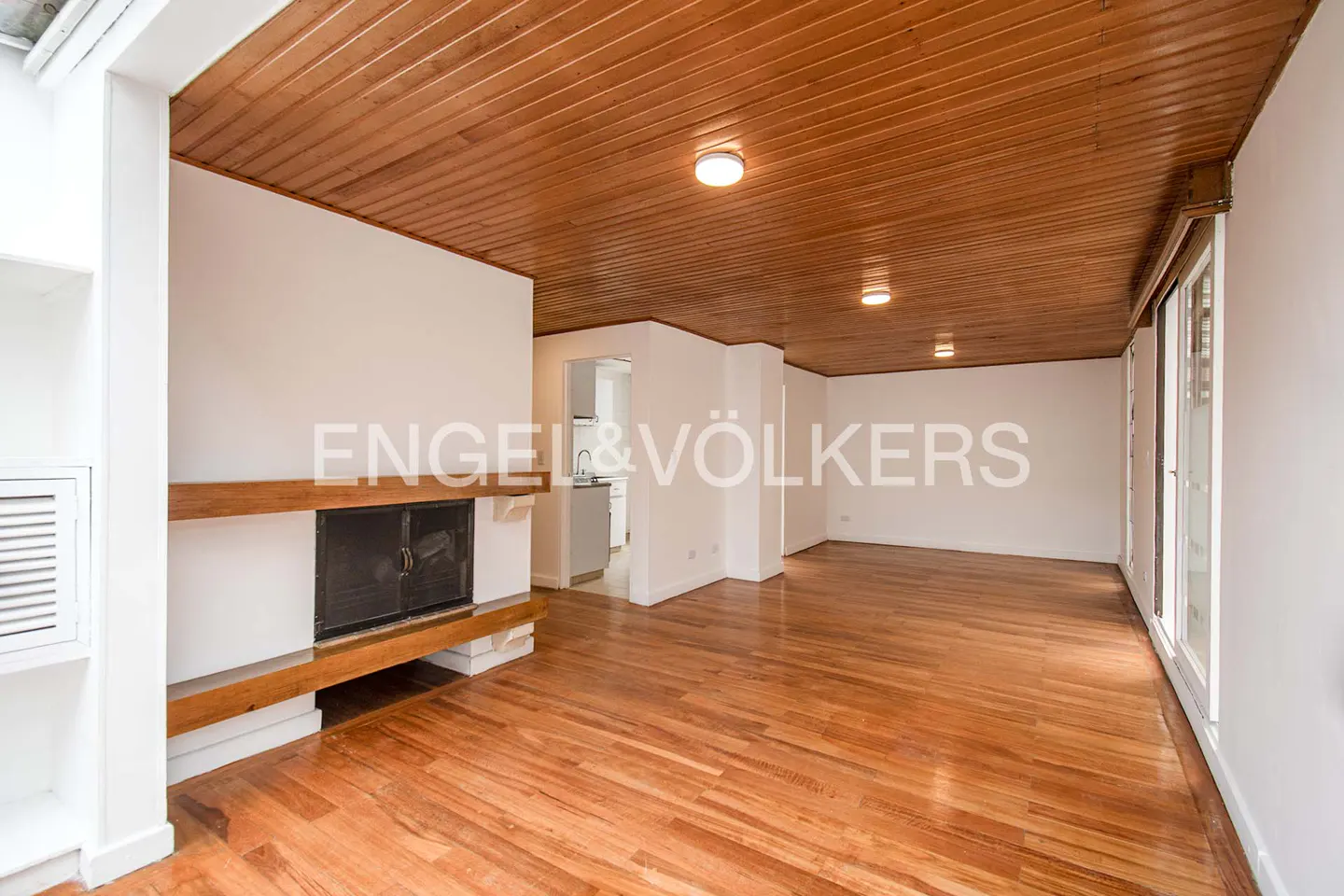 Spacious living room with wood-paneled ceiling, hardwood floors, a fireplace, and a view into a bright kitchen.