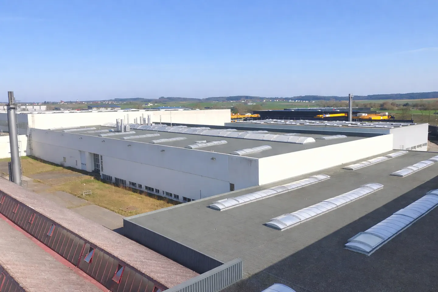Aerial view of a large, white industrial building with skylights on a gray roof under a clear blue sky.