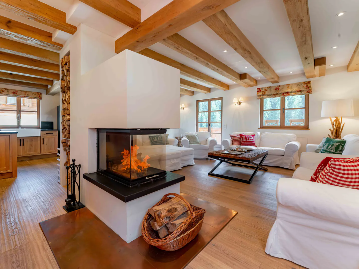 Bright living room with wood floors, exposed beams, and a modern fireplace with a basket of logs. White sofas and floral window treatments.