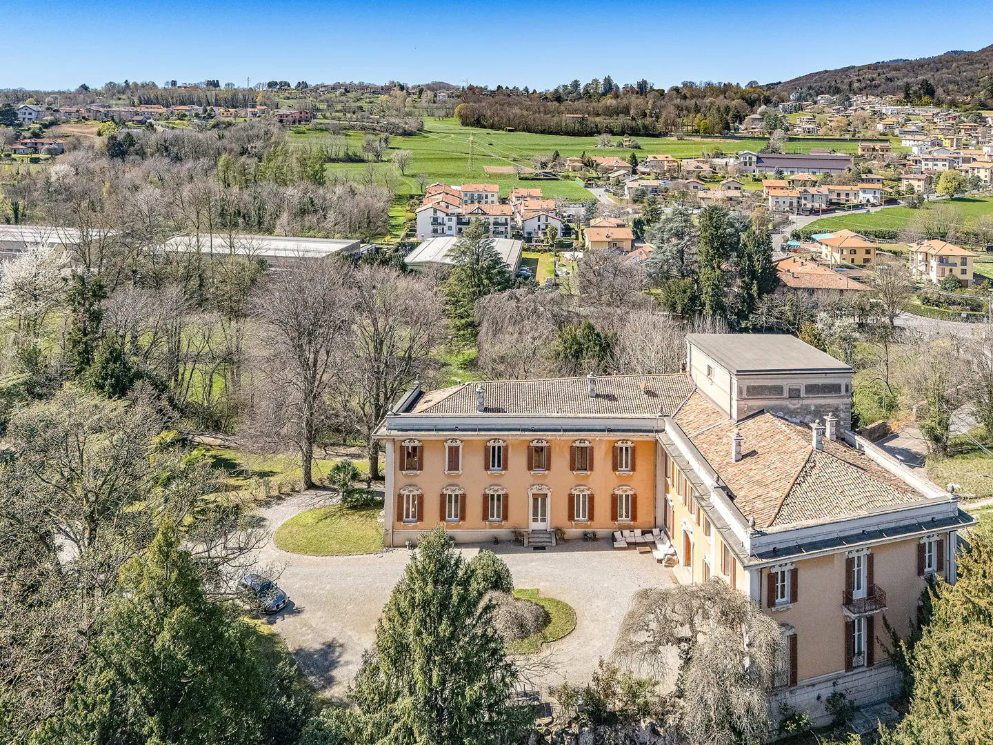 Aerial view of a large, peach-colored villa with brown shutters, surrounded by green trees and a gravel driveway. Rolling hills and a town are in the background.