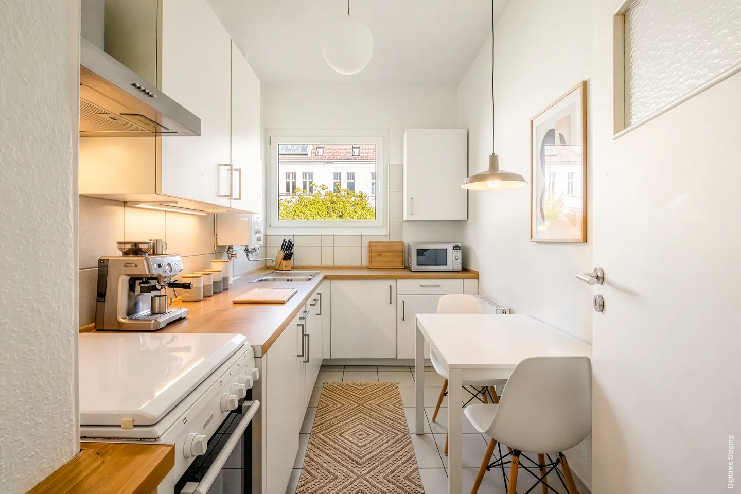 Bright, narrow kitchen with white cabinets, wood countertops, and a small dining table with two chairs. A window overlooks green trees.