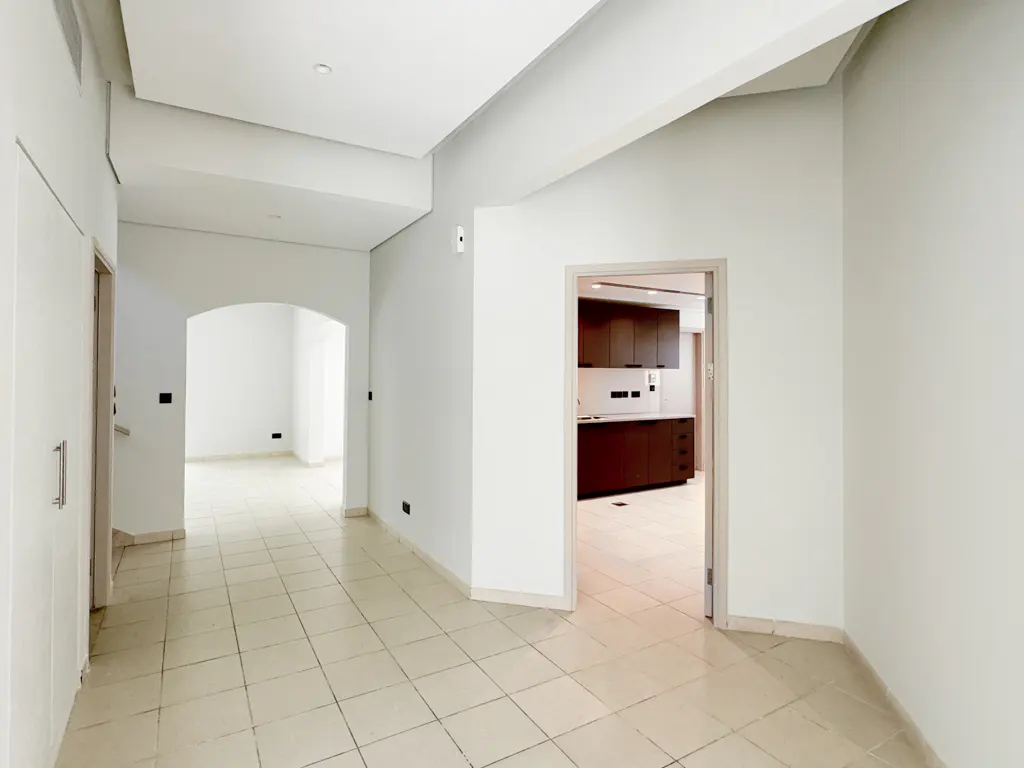 Bright, empty room with beige tile floor, white walls, and doorways to other rooms. Dark wood kitchen cabinets are visible through one doorway.