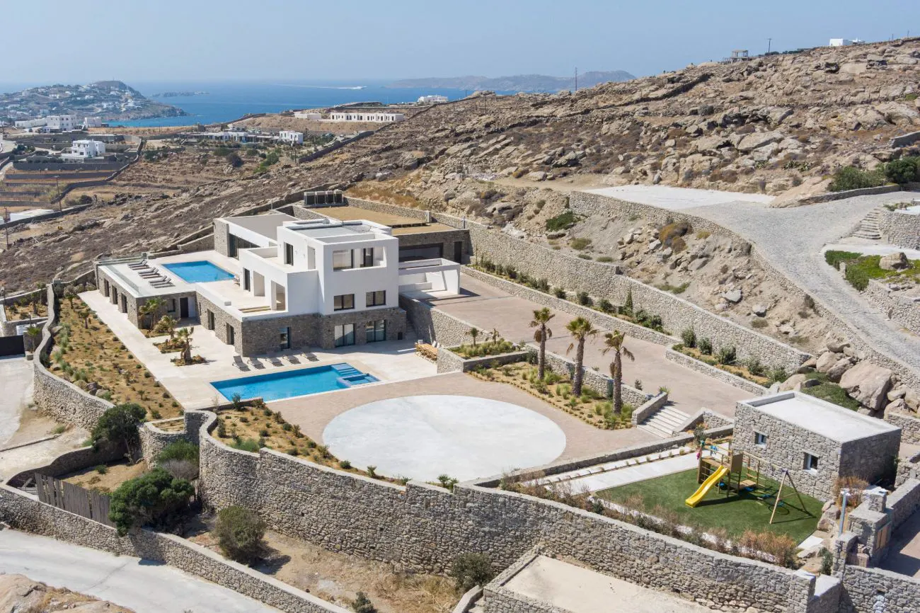 Aerial view of a modern white house with pools, palm trees, and a playground on a rocky hillside overlooking the ocean.