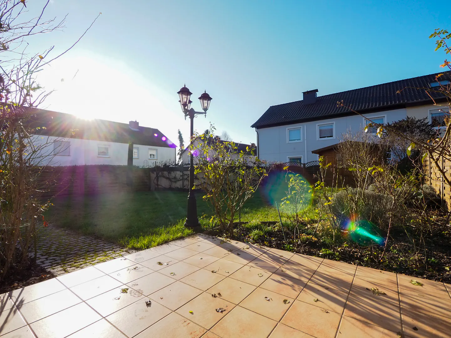 Sunny backyard with tile patio, green lawn, and a black lamppost. Houses are visible in the background under a clear blue sky.