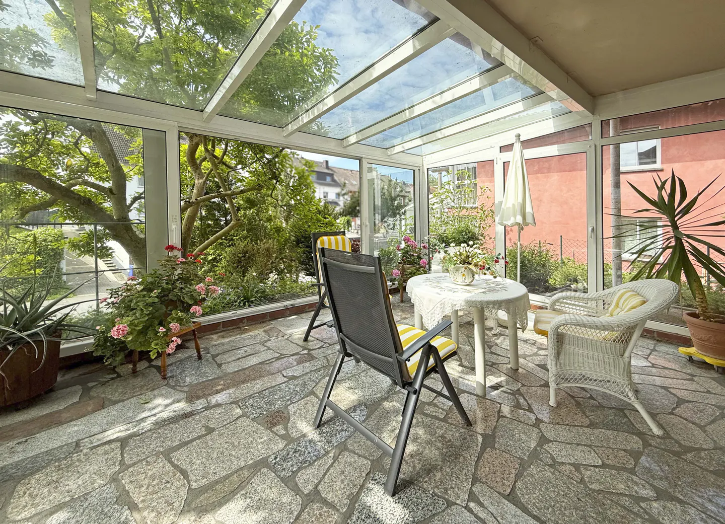 Sunroom with glass roof, stone floor, and garden view. Table, chairs, and plants create a cozy indoor-outdoor space.