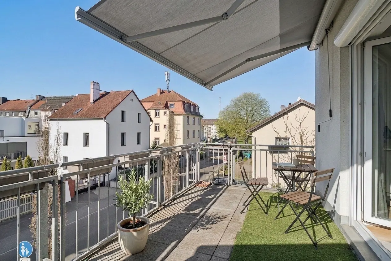 Balcony view with gray awning, metal railing, and potted plant. Table and chairs sit on artificial grass, with buildings in the background.