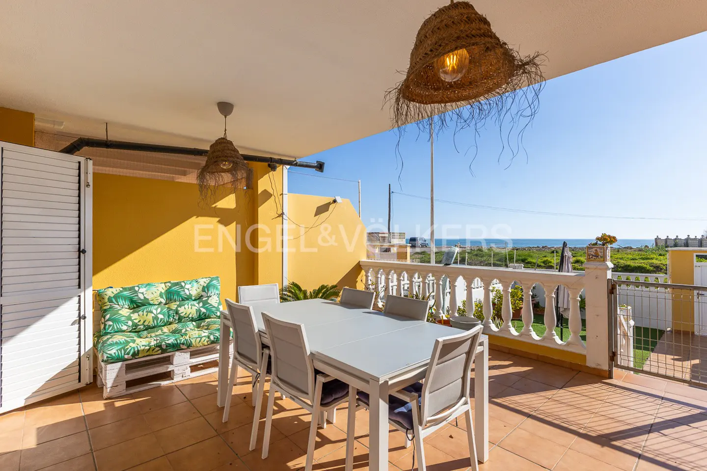 Covered patio with a white table, chairs, and a pallet bench with green cushions. Wicker lamps hang above, and the ocean is visible in the background.