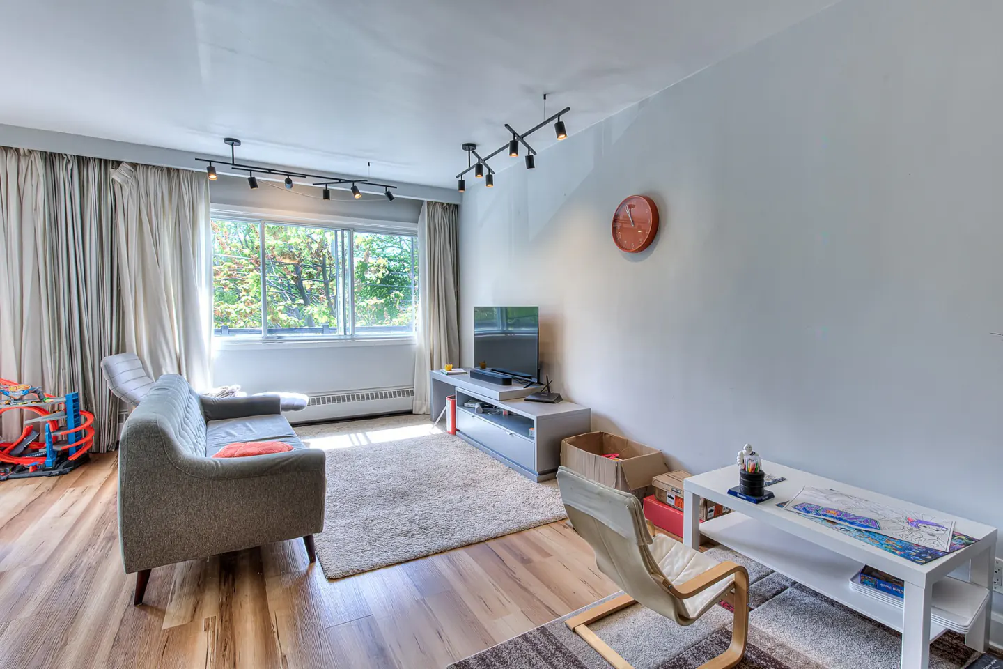 Living room with a gray sofa, TV, white walls, and wood floors. A large window shows trees outside. Track lighting illuminates the room.