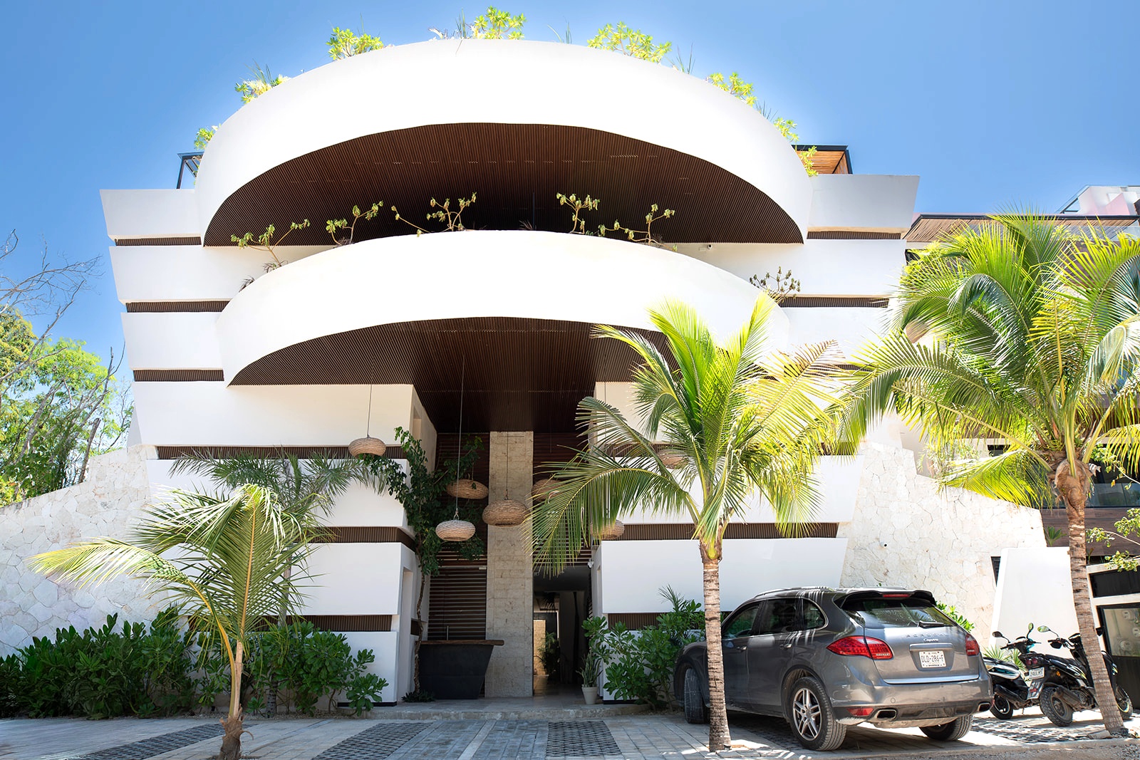 Modern white building with curved balconies and greenery, a gray car parked in front, and palm trees.