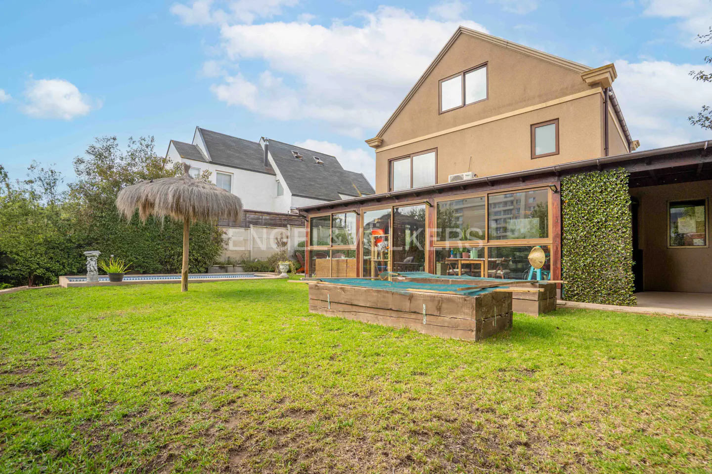 Backyard view of a tan two-story house with a glass-enclosed porch, a pool with a thatched umbrella, and a green lawn.
